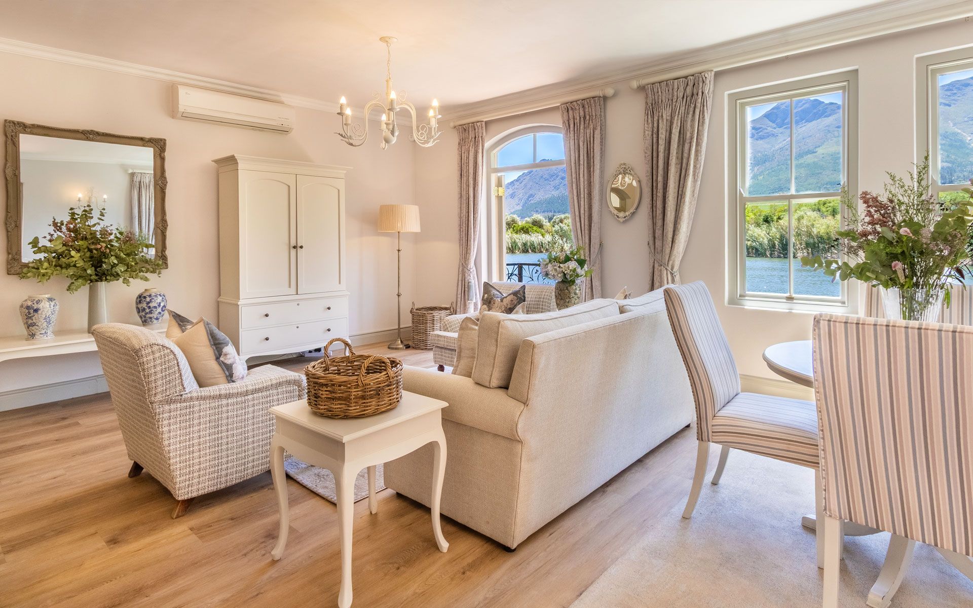 A beautifully decorated living room in Villa 11A, featuring plush sofas, a wicker basket, and elegant furnishings. Large windows offer stunning views of Patrysfontein Lake and the surrounding mountains, enhancing the serene atmosphere. A chandelier and floral arrangements add a touch of luxury.