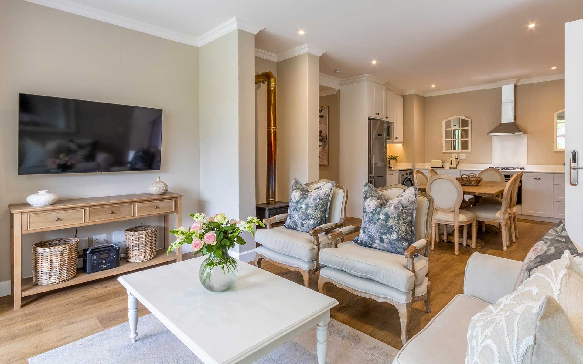 A cozy living area in Villa 56 features plush armchairs with floral cushions, a white coffee table with a flower vase, and a wall-mounted TV. The open kitchen includes modern appliances, a dining table, and soft lighting, creating a warm and inviting atmosphere.