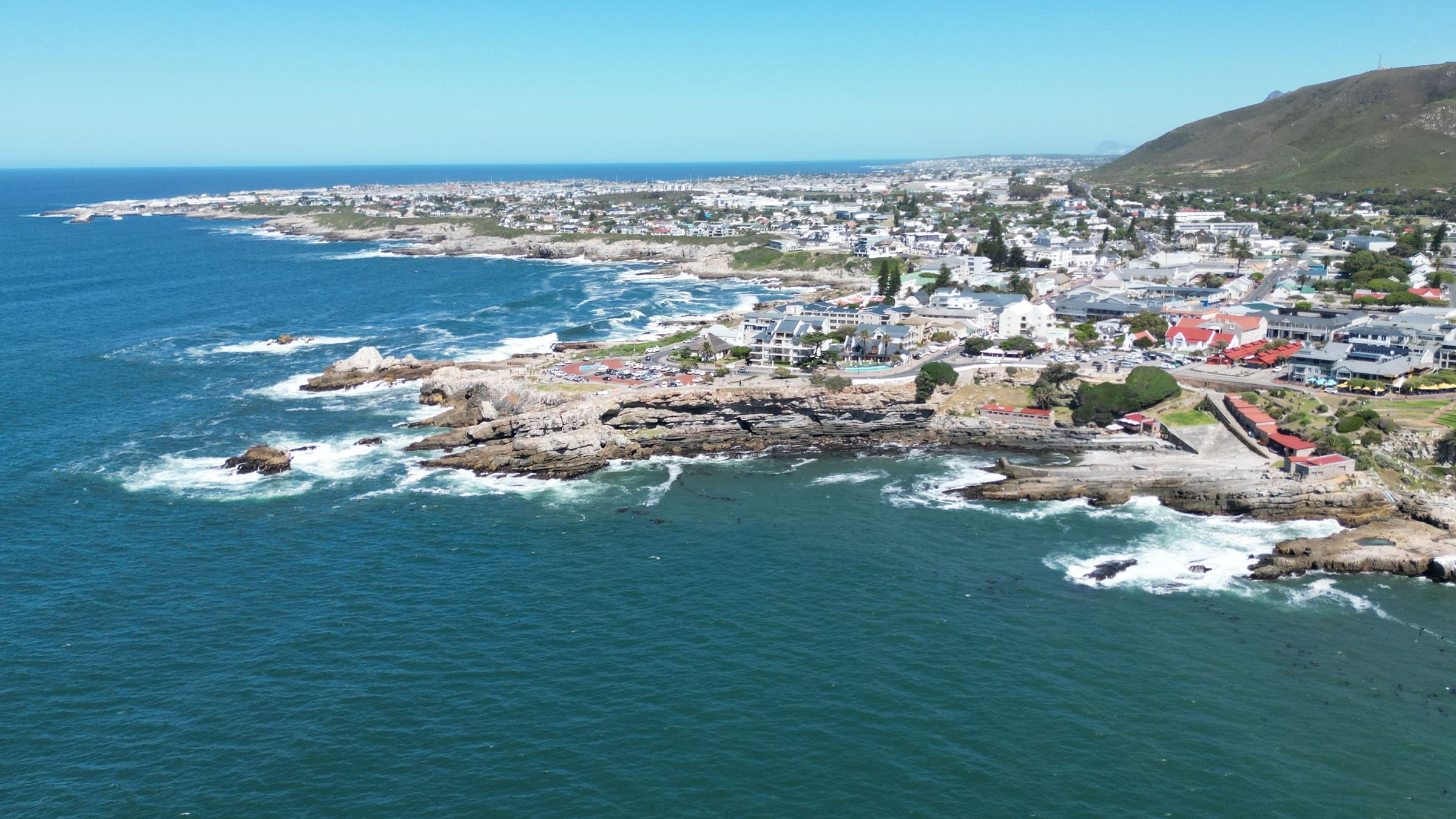 Aerial view of a coastal town with rocky shores and clear blue waters. The town is situated along the coastline with residential and commercial buildings visible. Waves crash against the rocks, and a hilly landscape is seen in the background under a clear sky.