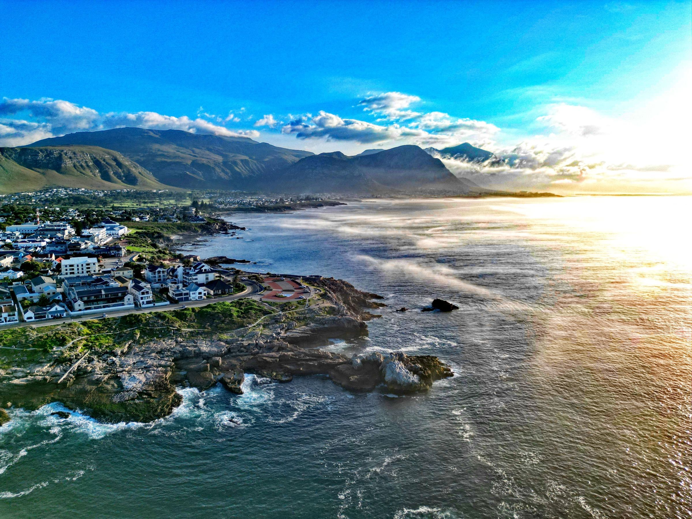 Aerial view of a coastal town with buildings near rocky shores and a calm sea reflecting the golden hues of sunrise. In the background, green hills and mountains are partially covered by clouds under a clear blue sky.