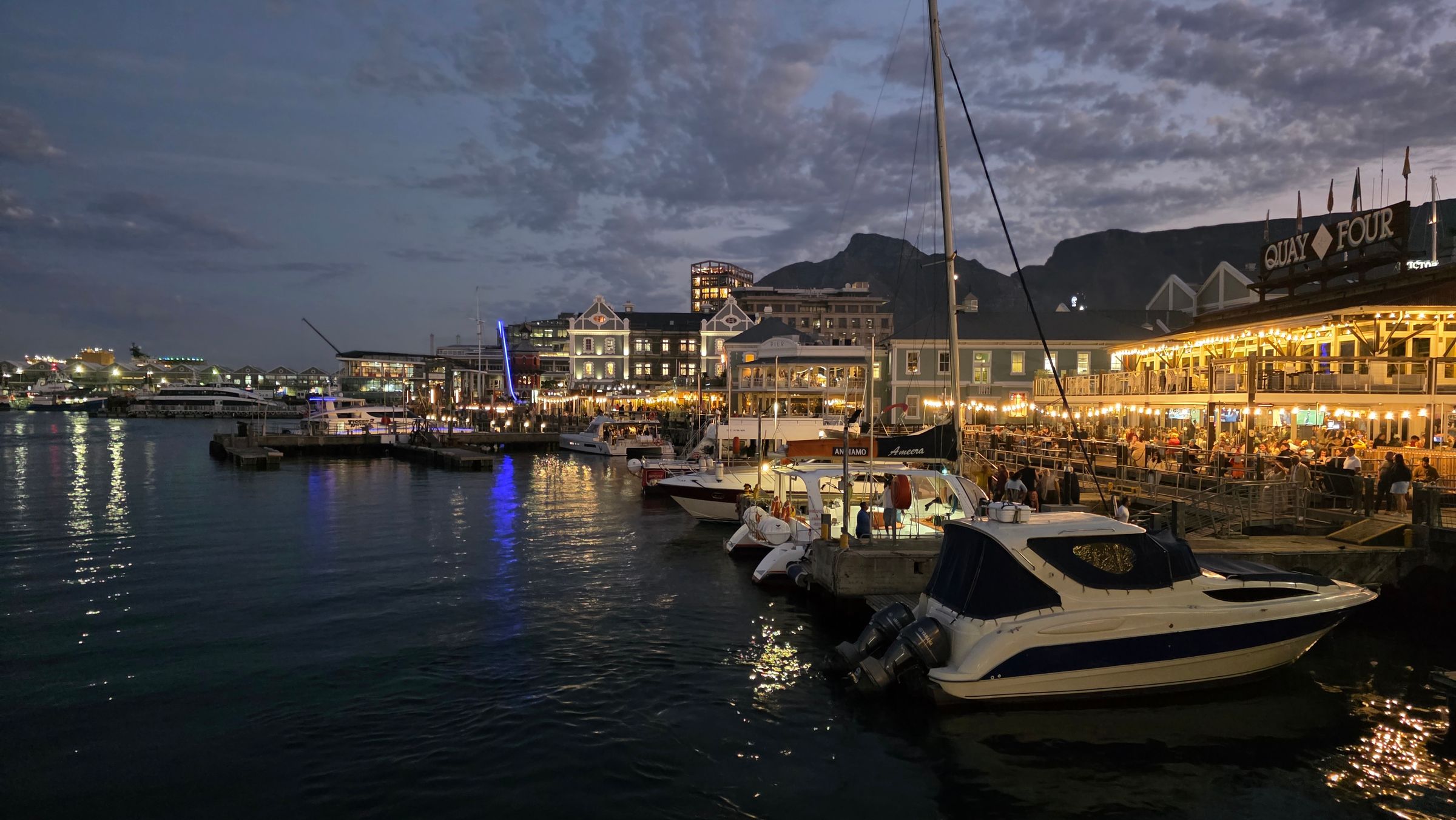 The V&A Waterfront in Cape Town at dusk, featuring illuminated boats docked along the harbor. Bustling restaurants and shops line the waterfront, with Table Mountain silhouetted against a twilight sky. The scene captures the lively mix of maritime heritage and modern vibrancy.