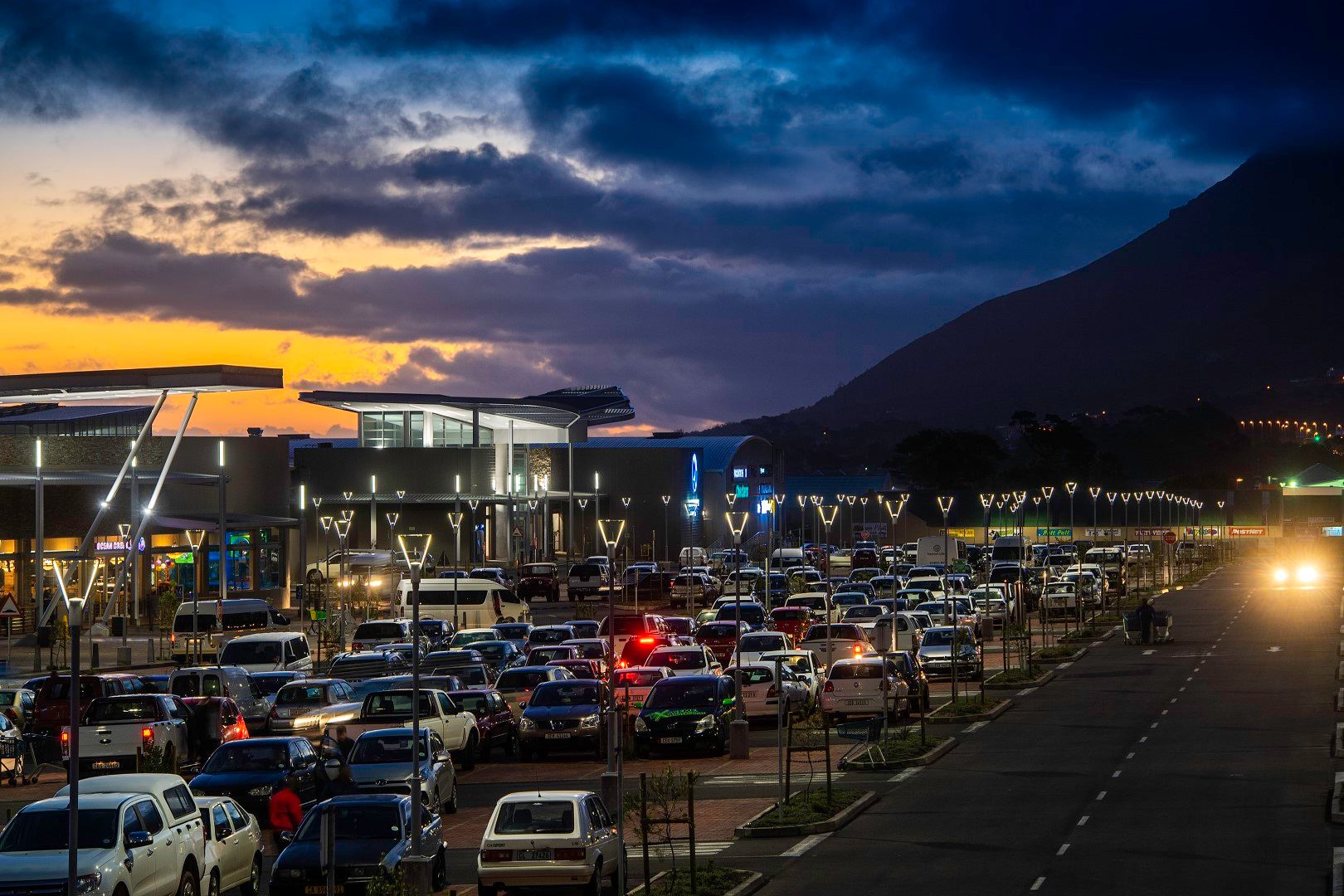 Twilight view of Whale Coast Mall with a bustling parking lot and illuminated storefronts. The modern architecture stands against a vibrant sunset, highlighting the mall's role as a central shopping hub on the Hermanus coastline, accessible to locals and visitors alike.
