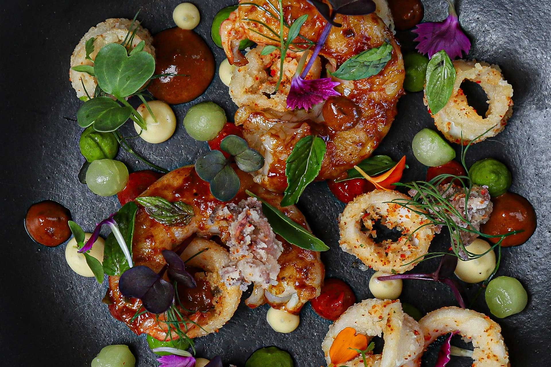 An artistic presentation of a gourmet seafood dish featuring fried calamari rings, shrimp, and colorful garnishes on a black plate. The dish includes vibrant greens, sauces, and edible flowers, creating a visually appealing culinary arrangement.