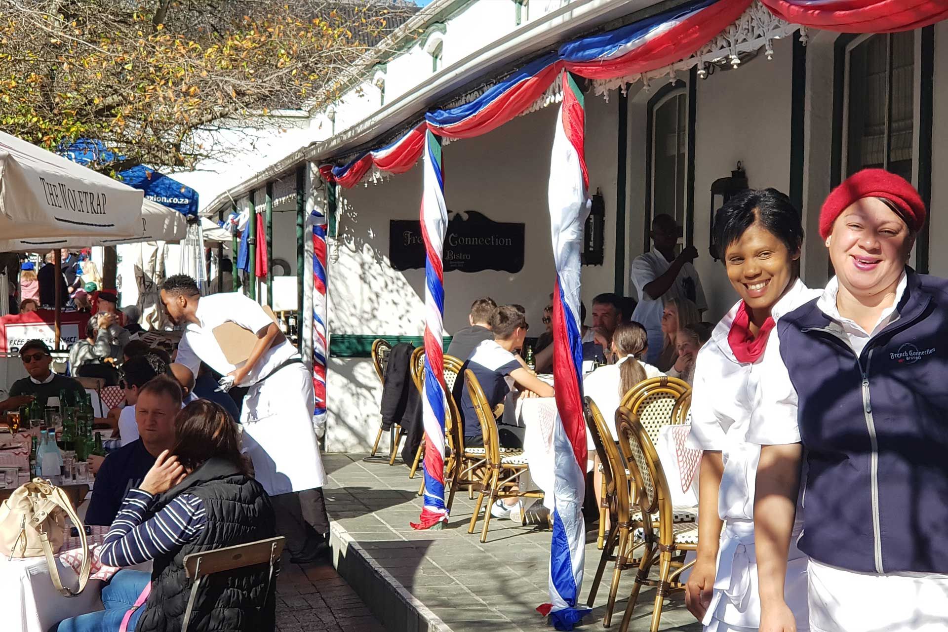 A vibrant outdoor dining scene at a bistro with red, white, and blue decorations. Two smiling staff members stand in the foreground, while patrons enjoy meals under umbrellas. The atmosphere is lively, with sunlight casting shadows on the cafe's porch and tables filled with people.