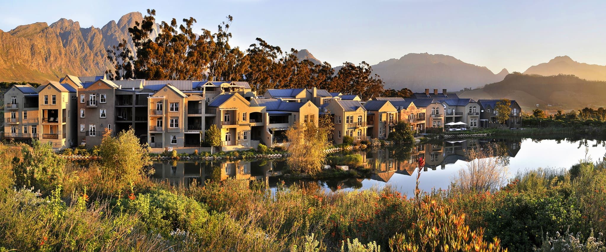 View of modern apartment buildings by a tranquil lake with mountains in the background, surrounded by lush greenery and bathed in warm sunlight, reflecting a peaceful and picturesque setting.