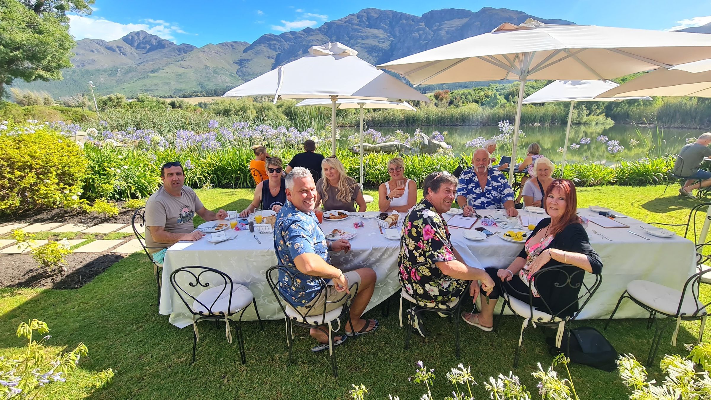 A group of people enjoying a meal at an outdoor table under umbrellas, set in a scenic landscape with a lake and mountains in the background. The table is elegantly set with white cloth and surrounded by lush greenery and flowering plants.