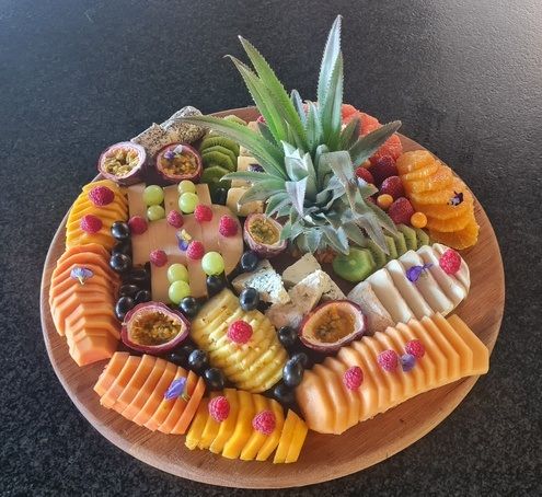 A circular wooden platter filled with an assortment of sliced fruits, including papaya, mango, kiwi, passion fruit, grapes, and blueberries. A decorative pineapple top is placed in the center, and edible flowers are sprinkled throughout for garnish.