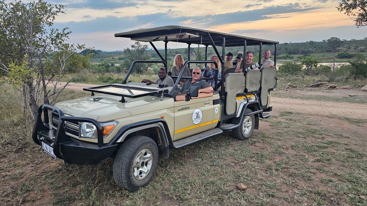 A group of people seated in an open safari jeep parked on a dirt path. The vehicle is beige with a logo on the side. The background features a grassy landscape with trees and a river under a cloudy sky. The passengers are smiling and enjoying the scene.