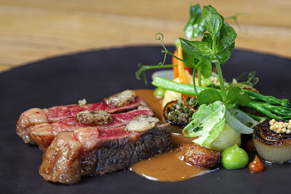 A beautifully plated gourmet dish featuring a perfectly cooked steak topped with mushrooms and surrounded by various vegetables including carrots, greens, and onions, all drizzled with a rich brown sauce on a dark plate.