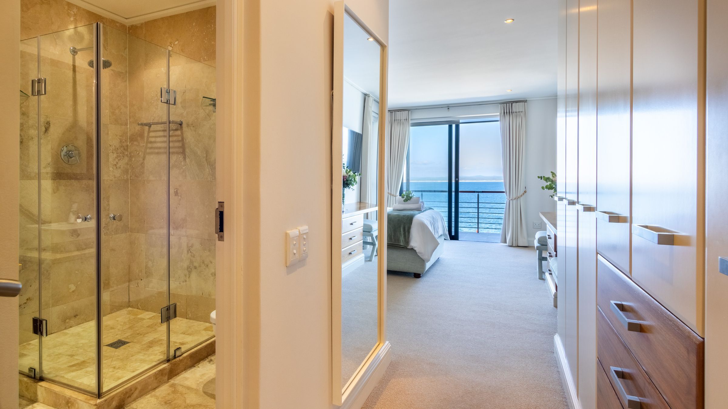 A modern bedroom with a glass shower enclosure to the left, featuring beige tiles. The room leads to a spacious area with a bed covered in white and green linens. Large windows offer a stunning ocean view. White wardrobes and a mirror enhance the space's elegance and brightness.