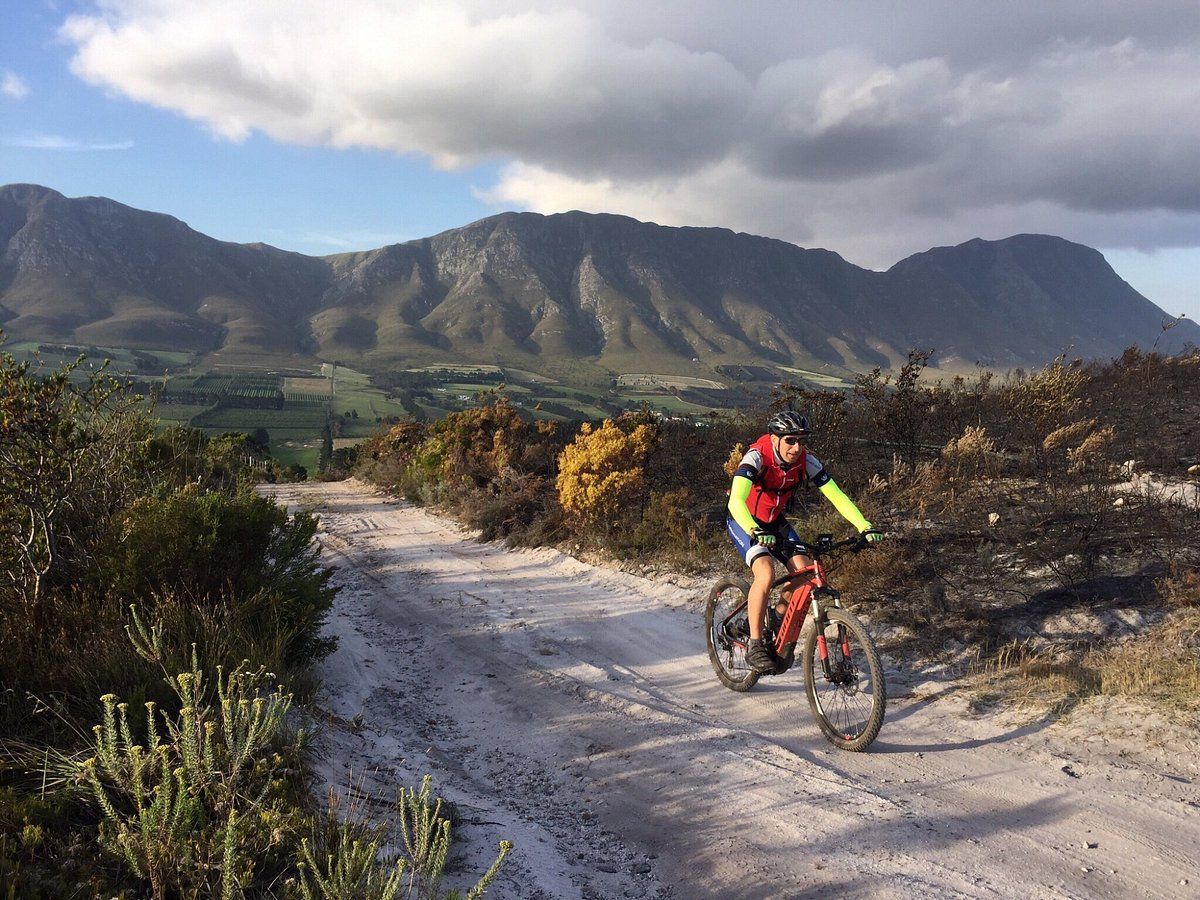 A cyclist rides on a dirt trail through a mountainous landscape. The rider wears a helmet, red vest, and neon sleeves. The background features lush greenery and rocky mountains under a partly cloudy sky, creating a picturesque outdoor scene.