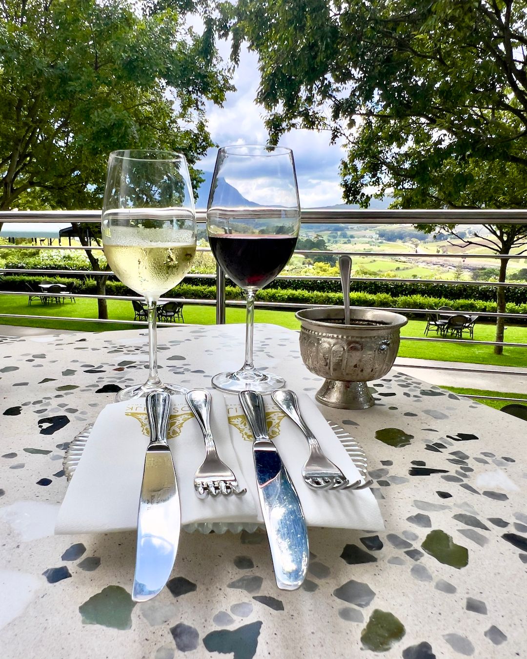 Two wine glasses, one with white and one with red wine, rest on a stone table with cutlery and a napkin. The backdrop features lush vineyards and a mountain under a partly cloudy sky, capturing the serene beauty of Hemel-en-Aarde wine valley in South Africa.
