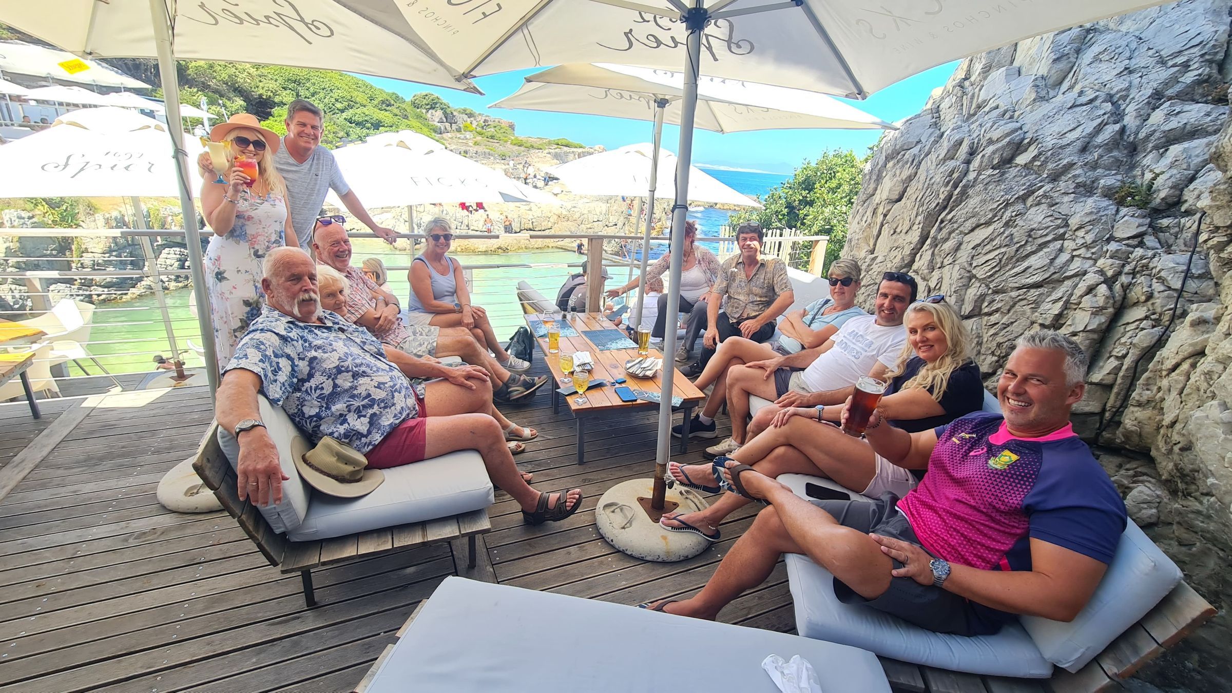 A group of people relaxing at an outdoor cafe with drinks, seated around a wooden table on cushioned benches under large umbrellas. The setting overlooks a scenic body of water and rocky cliffs, with greenery in the background and a clear blue sky overhead.