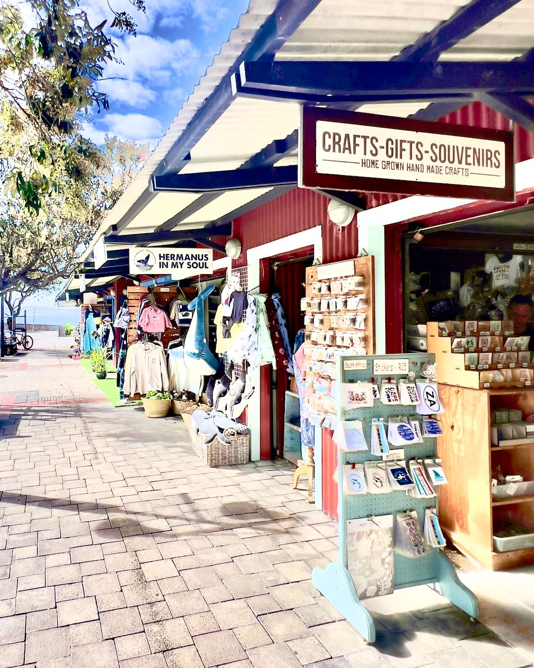 Market stalls in Hermanus displaying crafts, clothing, and souvenirs. Signs read 'Crafts-Gifts-Souvenirs' and 'Hermanus in My Soul'. Various items like postcards and apparel are visible, capturing the lively atmosphere and local artisans' contributions.