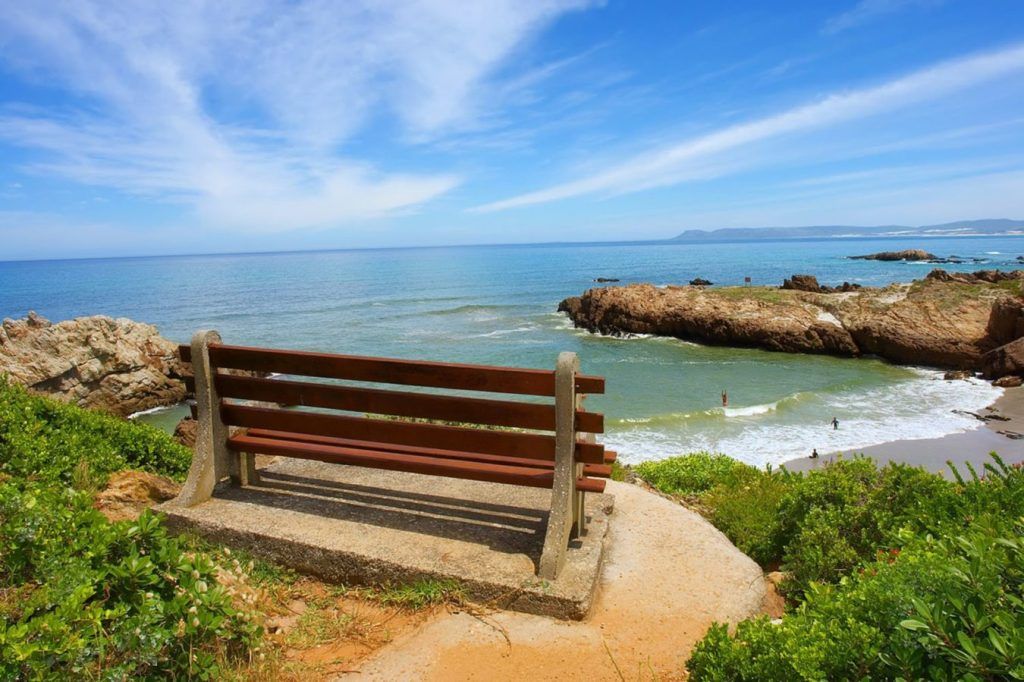 A wooden bench on a concrete slab overlooks a scenic ocean view with rocky cliffs and a sandy beach. The sky is clear with a few clouds, and the water is a mix of deep blue and turquoise hues. Greenery surrounds the bench, adding to the tranquil setting.