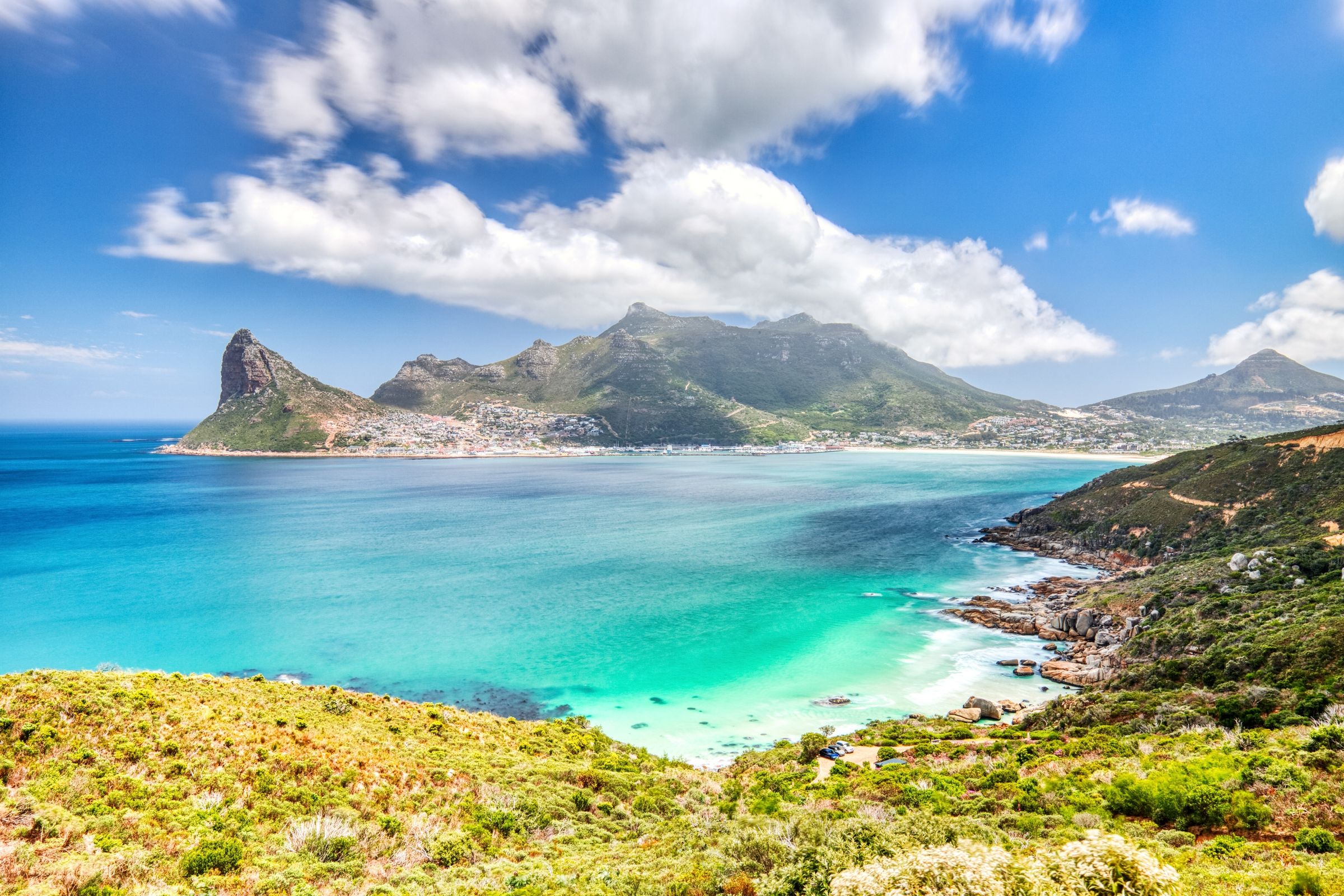 A stunning view of Hout Bay, showcasing the vibrant turquoise sea bordered by lush green mountains. The fishing village and natural harbor are nestled between the peaks, offering a picturesque escape with opportunities for seafood, seal watching, and coastal drives.