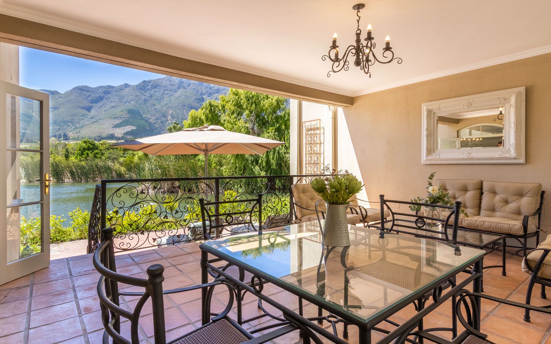 A charming lakeside terrace with a glass-topped dining table and cushioned seating. The space features elegant ironwork, a chandelier, and lush greenery. Open doors reveal a stunning view of the lake and mountains, offering a peaceful and picturesque setting.