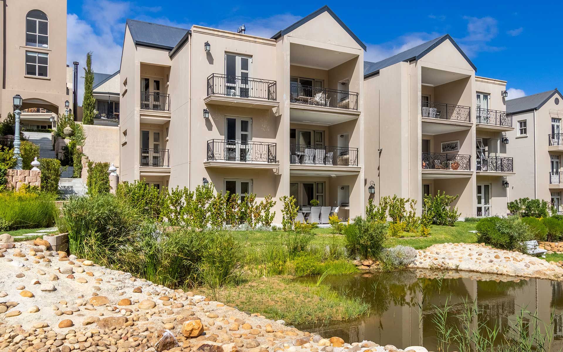View of a serene villa complex featuring multiple levels with balconies. Set against a backdrop of lush greenery and a tranquil pond, the architecture blends modern design with natural beauty, offering a peaceful retreat by the riverside.