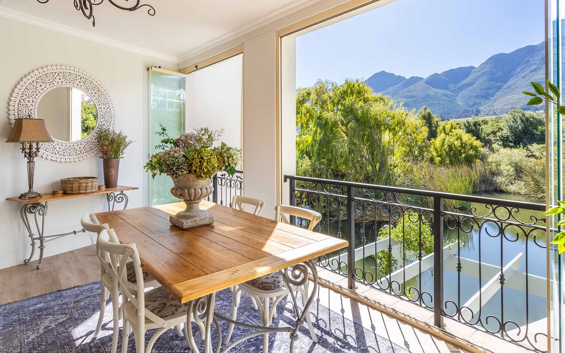 A rustic wooden dining table with elegant chairs sits on a balcony overlooking a serene lake, surrounded by lush greenery and mountains. A decorative mirror and lamp adorn a nearby console table, enhancing the villa's charm and tranquility.