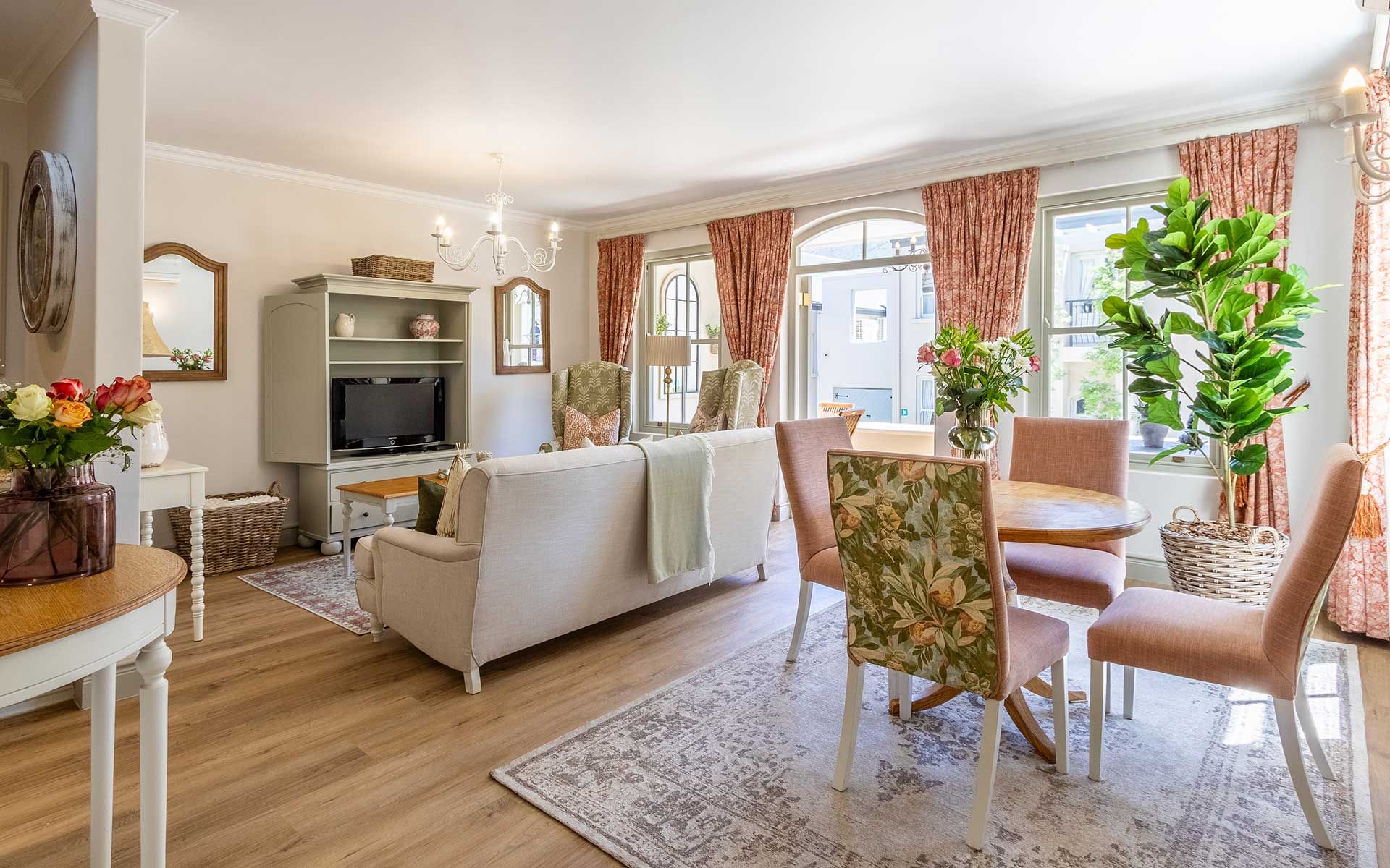 A bright, airy living space in Villa 27 featuring a comfortable sofa, floral-patterned chairs, and a wooden dining table. Large windows with pink curtains allow natural light, enhancing the cozy, contemporary decor. Potted plants and a TV on a light green cabinet complete the setting.