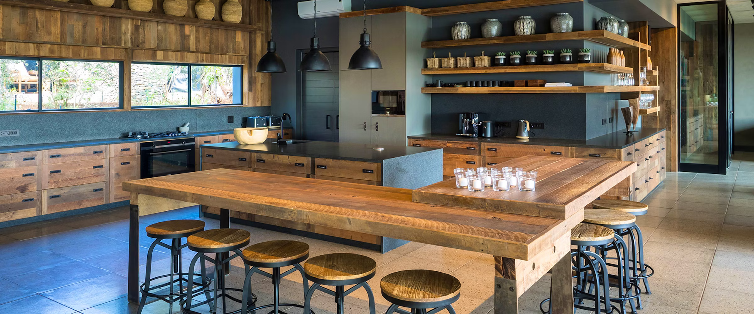 A modern kitchen with rustic wooden elements at Kruger Safari Lodge. Features include a large wooden table, bar stools, open shelving with decorative items, and contemporary lighting fixtures, creating a cozy yet upscale ambiance for luxury safari living.