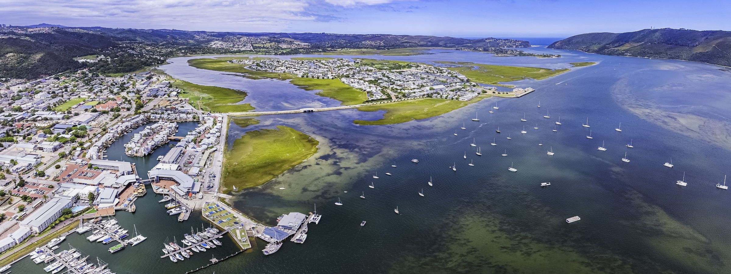 A panoramic aerial view of a coastal town with a marina filled with boats, surrounded by lush green landscapes and waterways. The town is nestled between rolling hills and the sea, with a bridge connecting different parts of the land. The sky is clear with a few clouds.