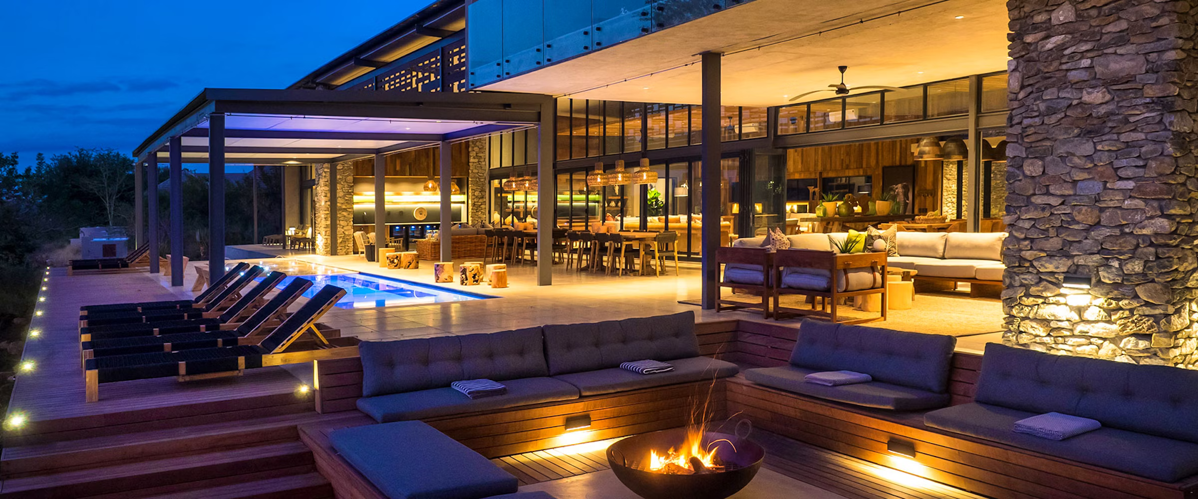 A modern terrace at dusk featuring a cozy fire pit surrounded by plush seating. The area includes a sleek pool, lined with wooden deck chairs, and a spacious open-concept interior with large glass windows and stone accents, warmly lit for a relaxing ambiance.