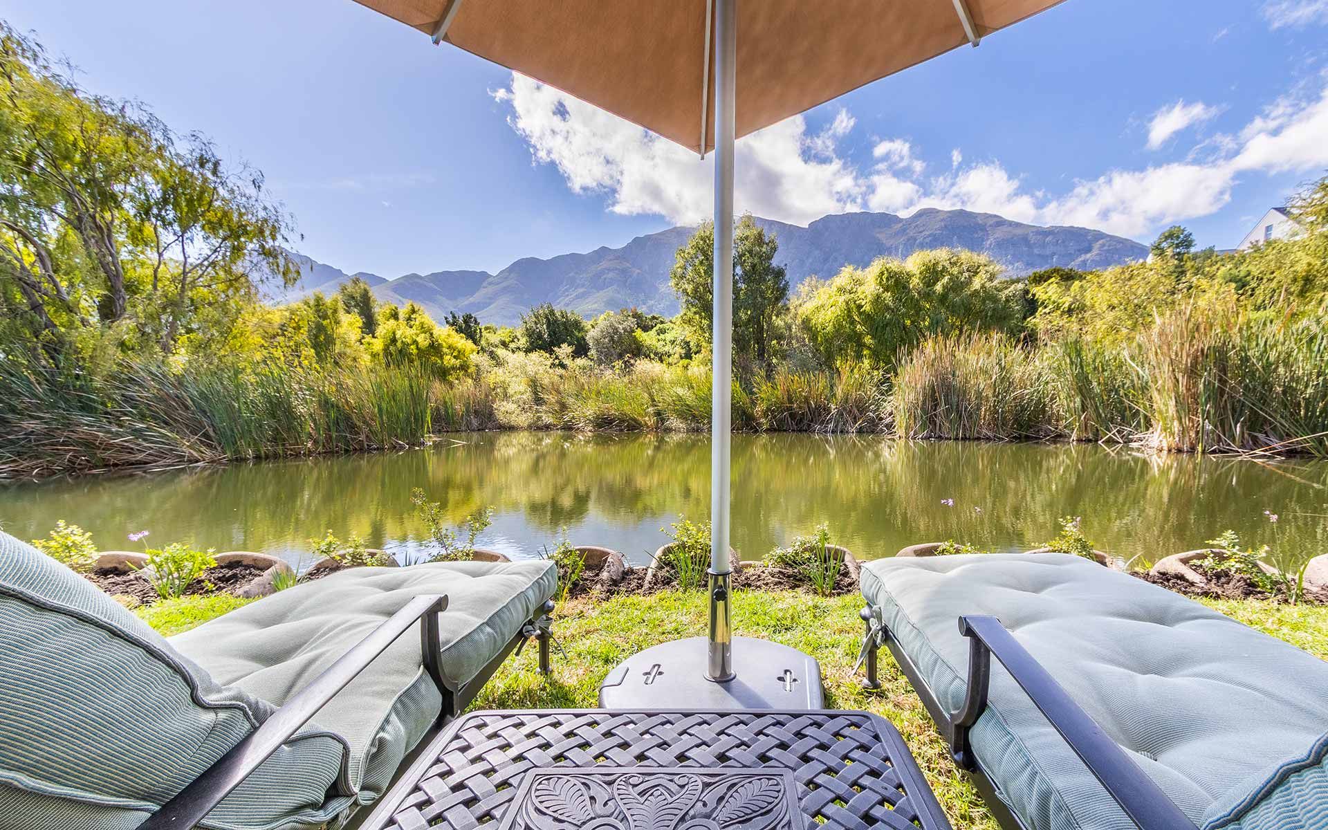 View from a private garden at Villa 31, featuring two loungers under an umbrella overlooking a serene lake and lush greenery. The backdrop includes the majestic Mont Rochelle mountains under a blue sky, offering a tranquil setting for relaxation and enjoyment.