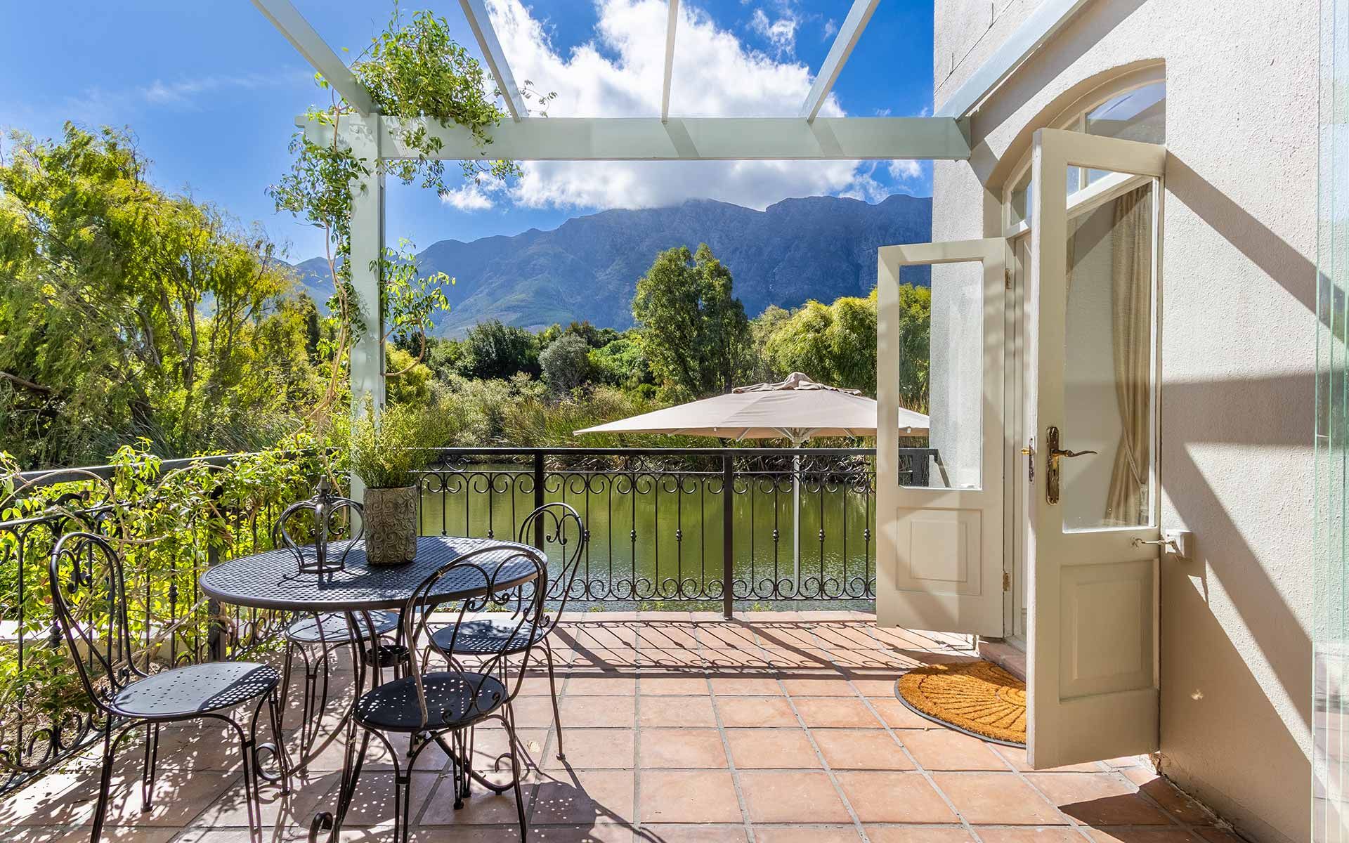 A charming outdoor terrace with a wrought iron table and chairs overlooks a tranquil lake, framed by lush greenery and majestic mountains under a clear blue sky. The open doorway invites natural light, enhancing the peaceful ambiance of this scenic retreat.