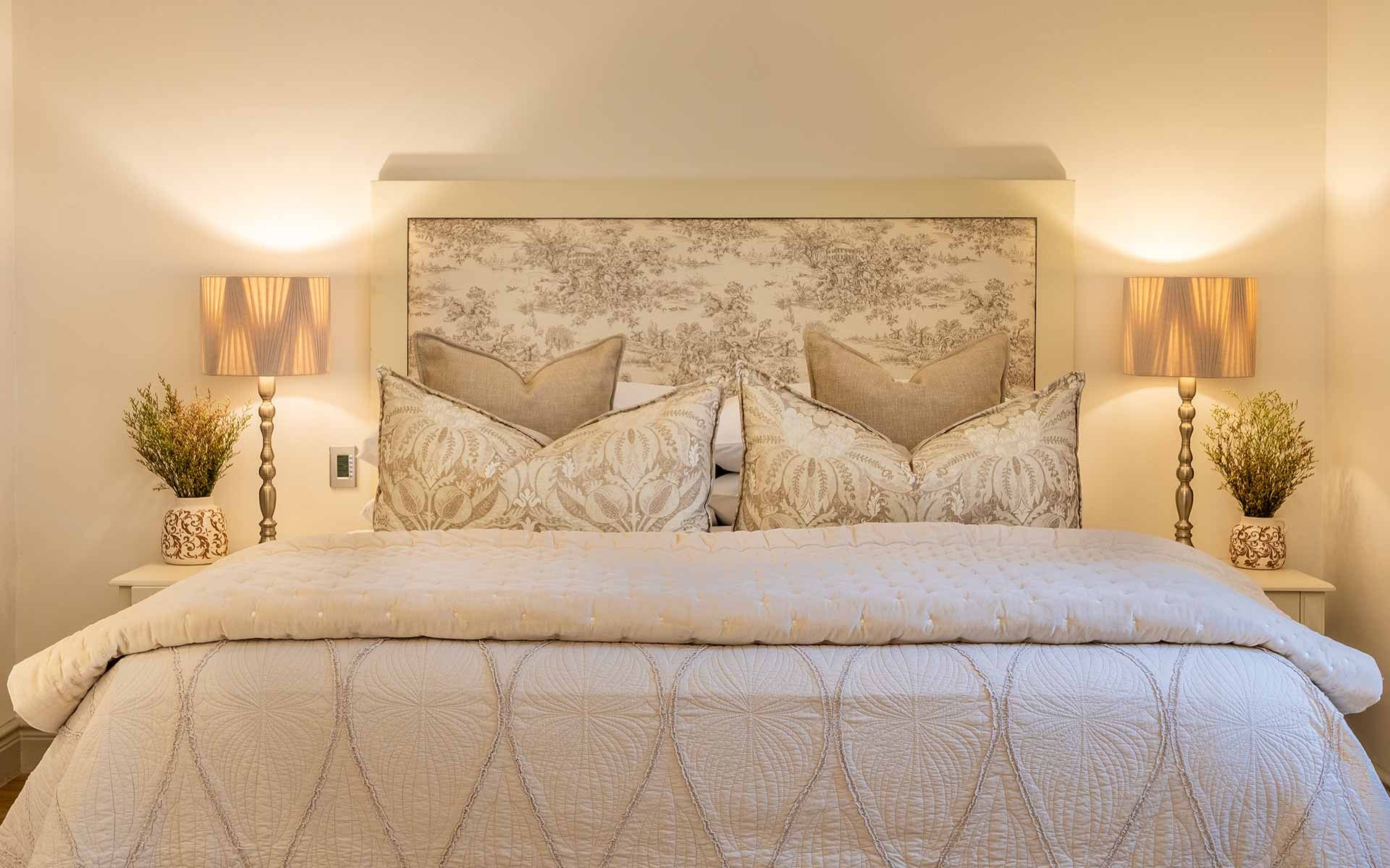 An elegant bedroom with a plush king-size bed adorned with patterned pillows and a textured quilt. Soft lighting from matching lamps on either side enhances the cozy atmosphere, complemented by decorative potted plants and a detailed headboard, creating an inviting retreat.