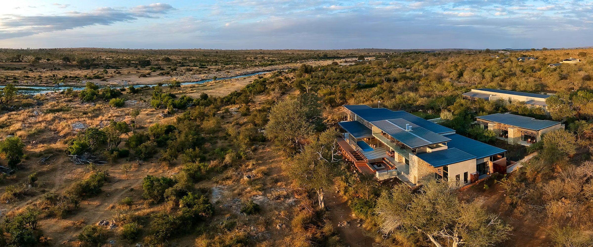 Aerial view of a modern lodge nestled in the African bush. The building features a sleek design with large windows and blue roofs, surrounded by sparse vegetation and a distant river under a partly cloudy sky.