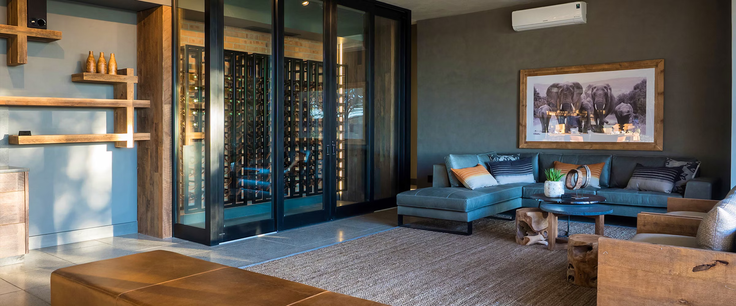 A stylish lounge featuring a sectional sofa, rustic wood accents, and a prominent wine display. The room is illuminated with warm lighting and decorated with wildlife art, creating a cozy and inviting atmosphere.