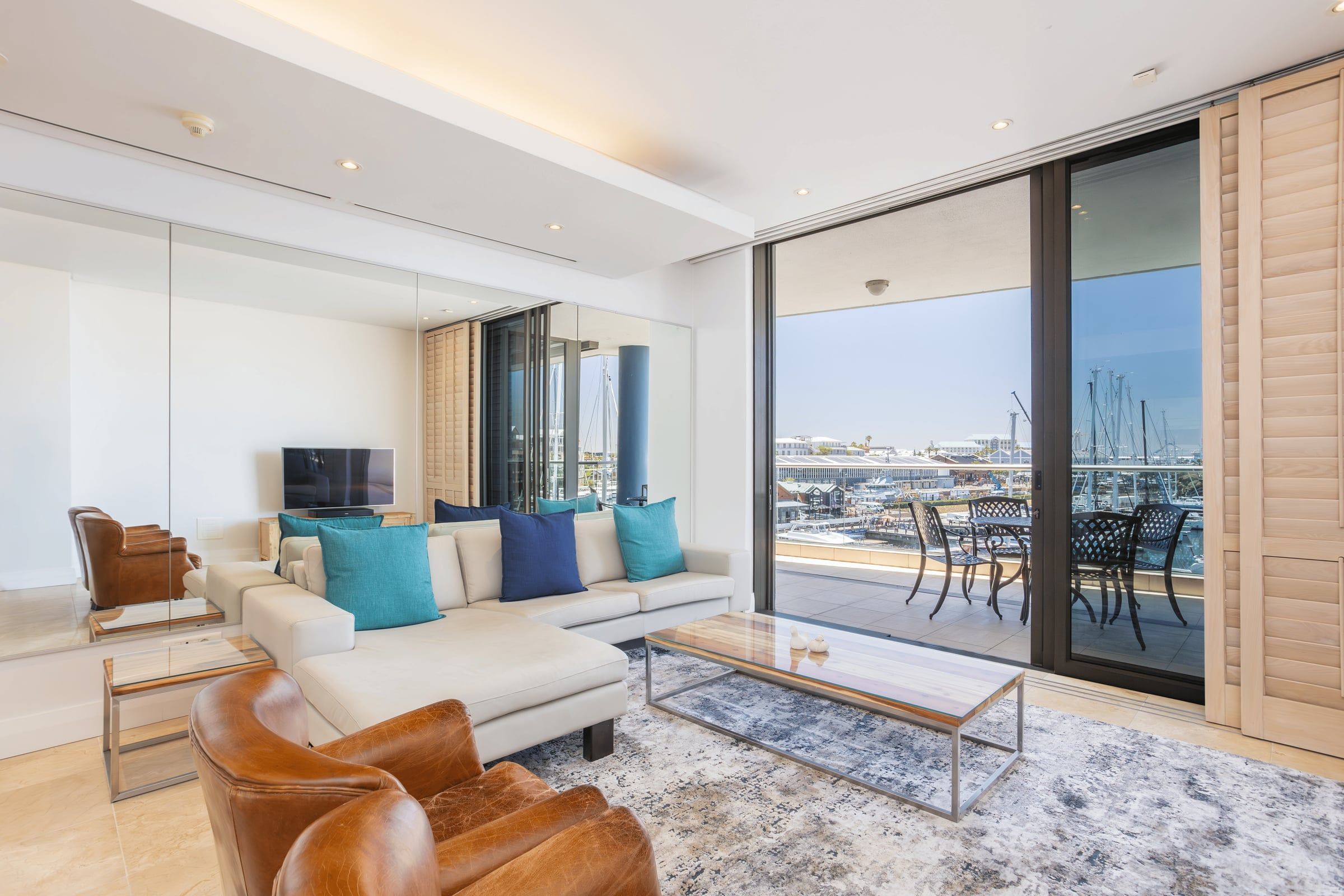 A modern living room with a beige sectional sofa adorned with blue and teal cushions, a leather armchair, and a glass coffee table. Large sliding doors open to a balcony with a marina view, featuring outdoor seating and visible sailboats. A TV is mounted on the wall.