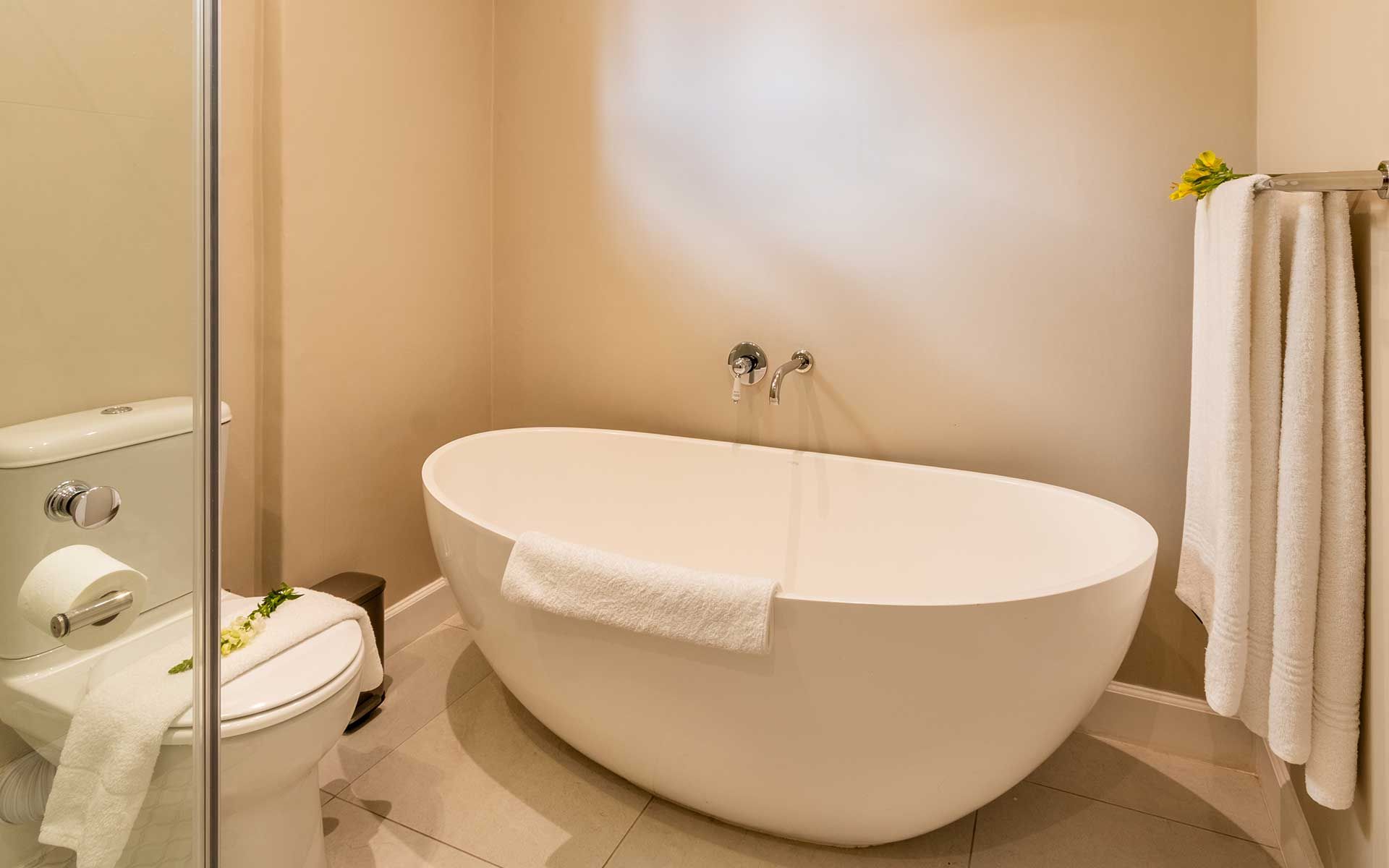 A spacious, modern bathroom features a sleek white freestanding bathtub with a towel draped over the edge. Nearby, a toilet is topped with a folded towel and a decorative flower. Soft lighting and neutral tones create a calm, inviting ambiance.