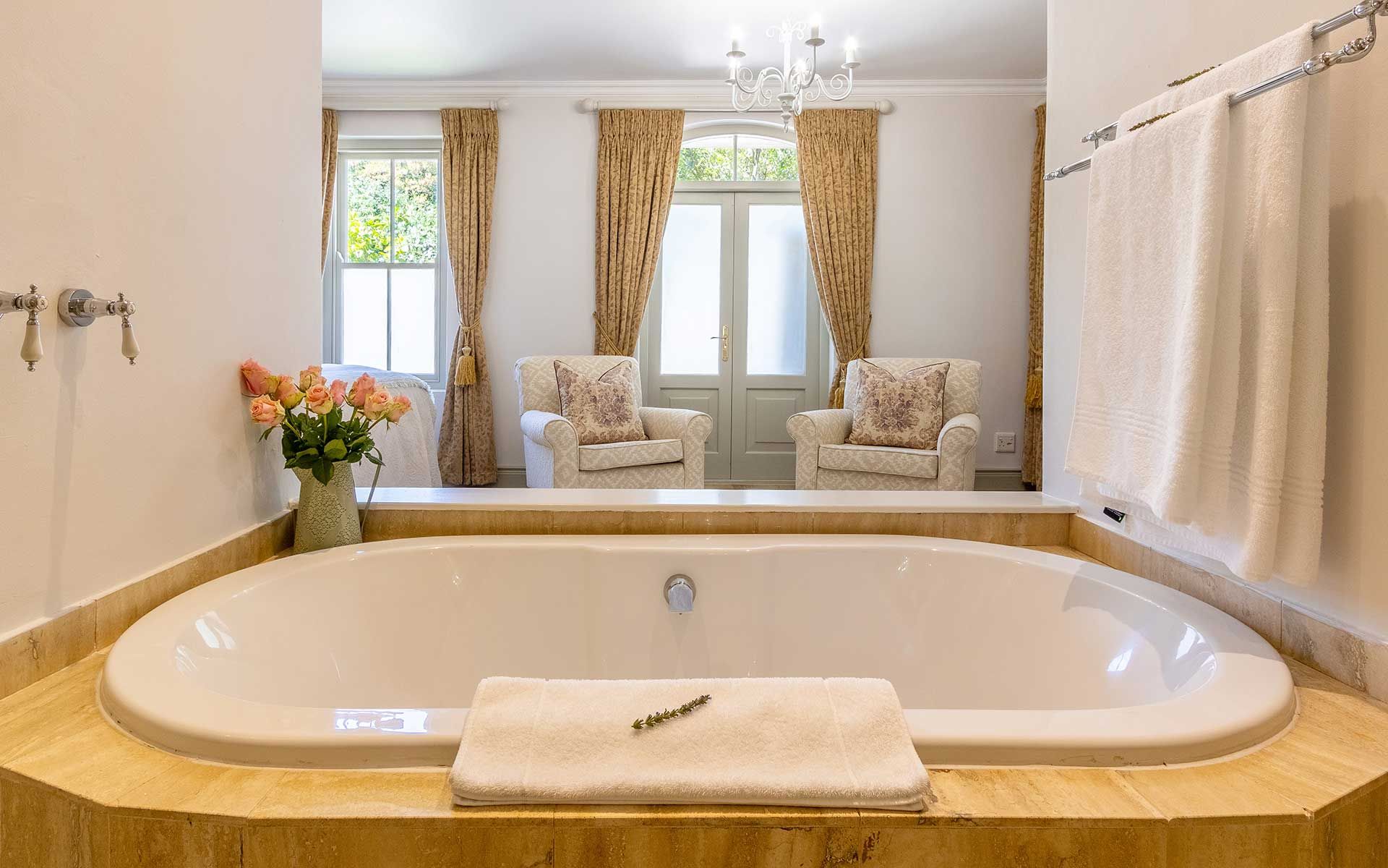 Elegant bathroom with a large soaking tub, adorned with fresh flowers and plush towels. Sunlight filters through large windows with beige curtains, revealing a cozy seating area with armchairs and a chandelier, creating a serene and luxurious atmosphere in Villa 27.