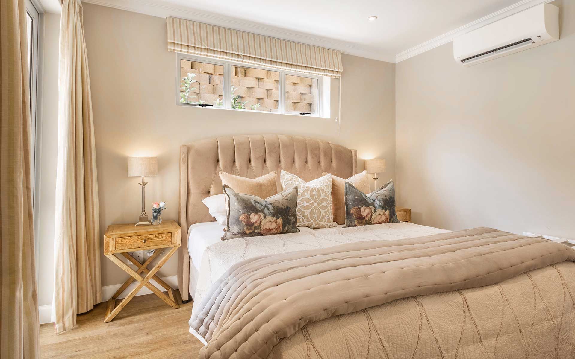 A cozy bedroom in Villa 56 featuring a plush king-size bed with floral pillows and a taupe quilt. The room is elegantly decorated with wooden bedside tables, soft lighting, and large windows with striped curtains, creating a serene and inviting atmosphere.