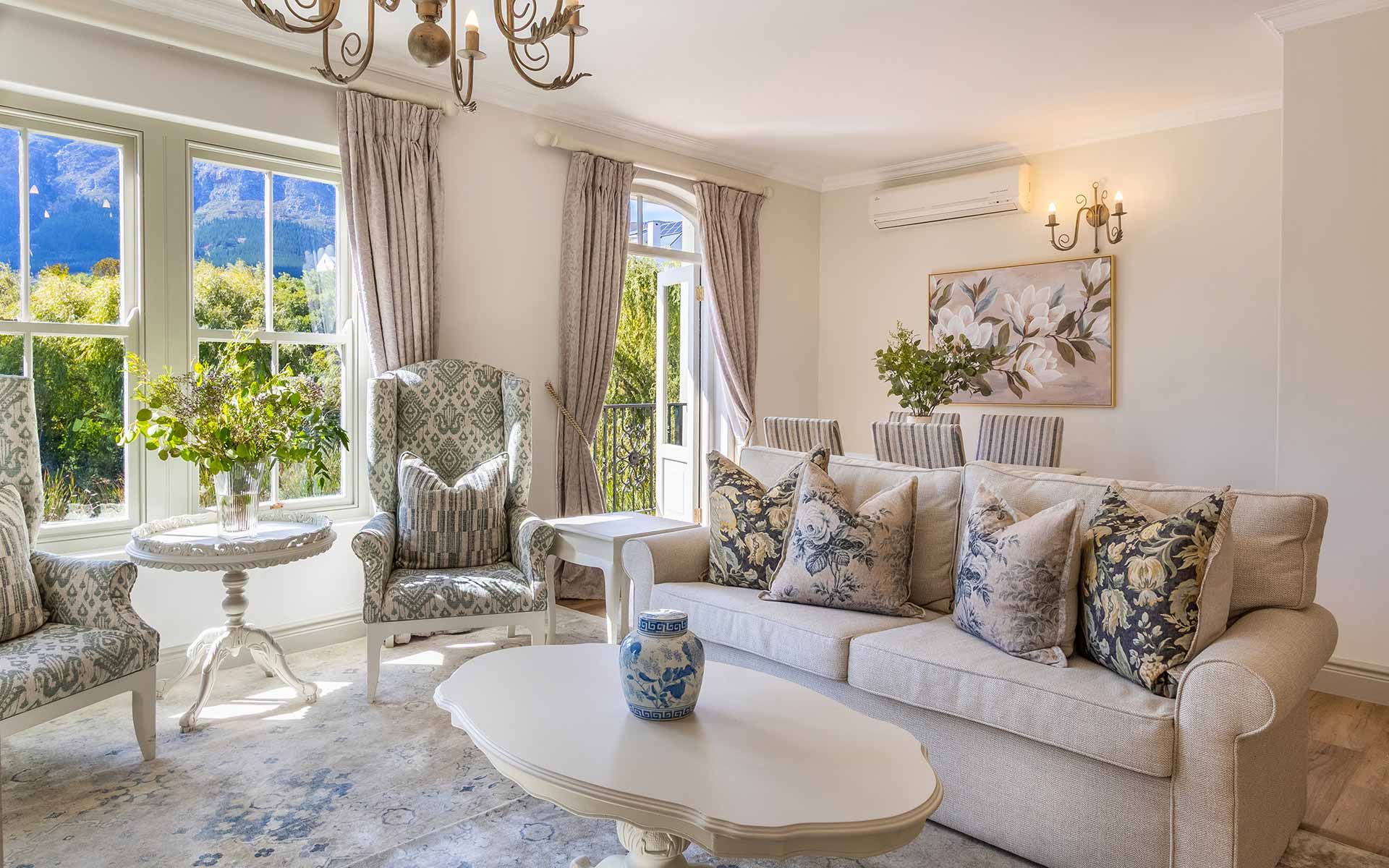 A bright living room in Villa 31 with elegant floral decor, plush seating, and large windows offering stunning views of nature. The room features a chic chandelier, a white coffee table with a decorative vase, and is softly lit by wall sconces, providing a cozy, upscale ambiance.