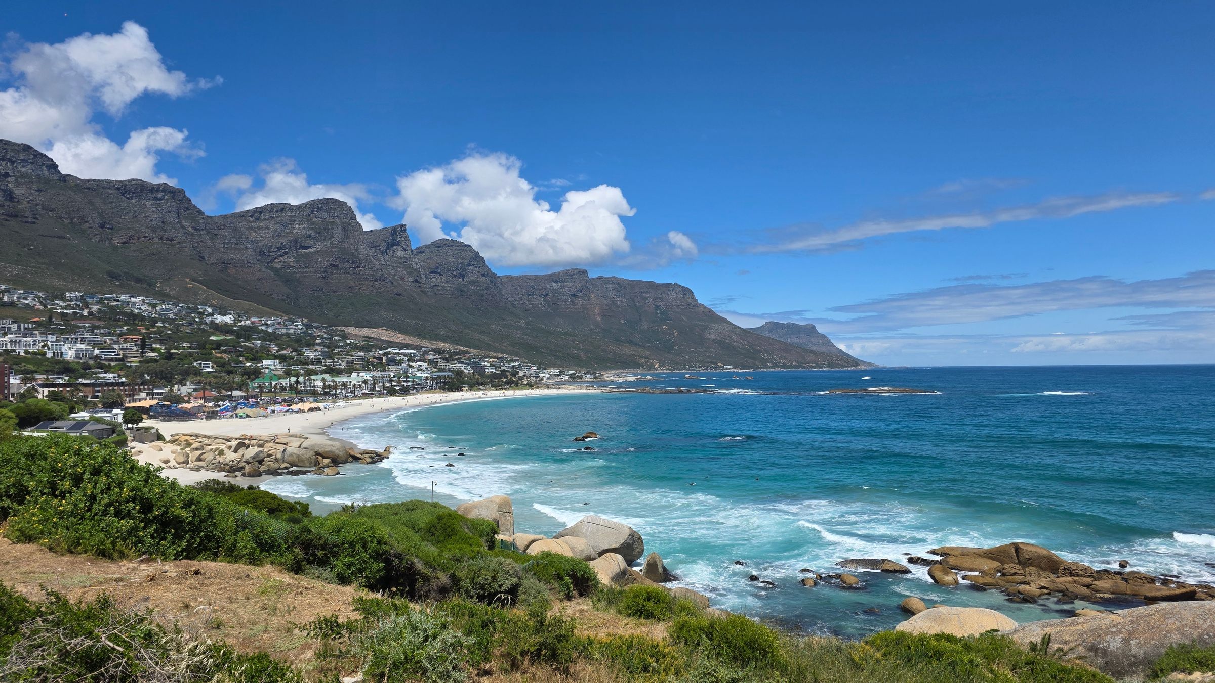 Scenic view of Cape Town's coastline featuring pristine white sands and turquoise Atlantic waters. The dramatic Twelve Apostles mountain range looms in the background, contrasting with the vibrant cityscape nestled along the shore. A clear blue sky enhances the natural beauty.