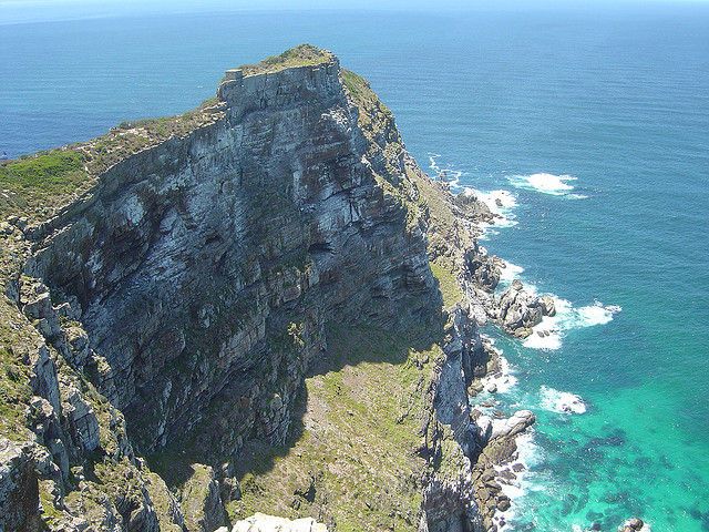 A striking view of the towering cliffs at Cape Point Nature Reserve, where rugged rock formations meet the vibrant blue waters of the ocean. The scene captures the dramatic convergence of the Atlantic and Indian Oceans, highlighting the raw natural beauty and power of the landscape.