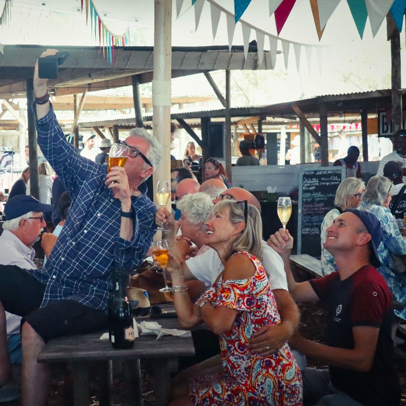 People taking a selfie while enjoying drinks at a lively market. The atmosphere is festive with colorful bunting overhead and a rustic setting, capturing a joyful community moment.