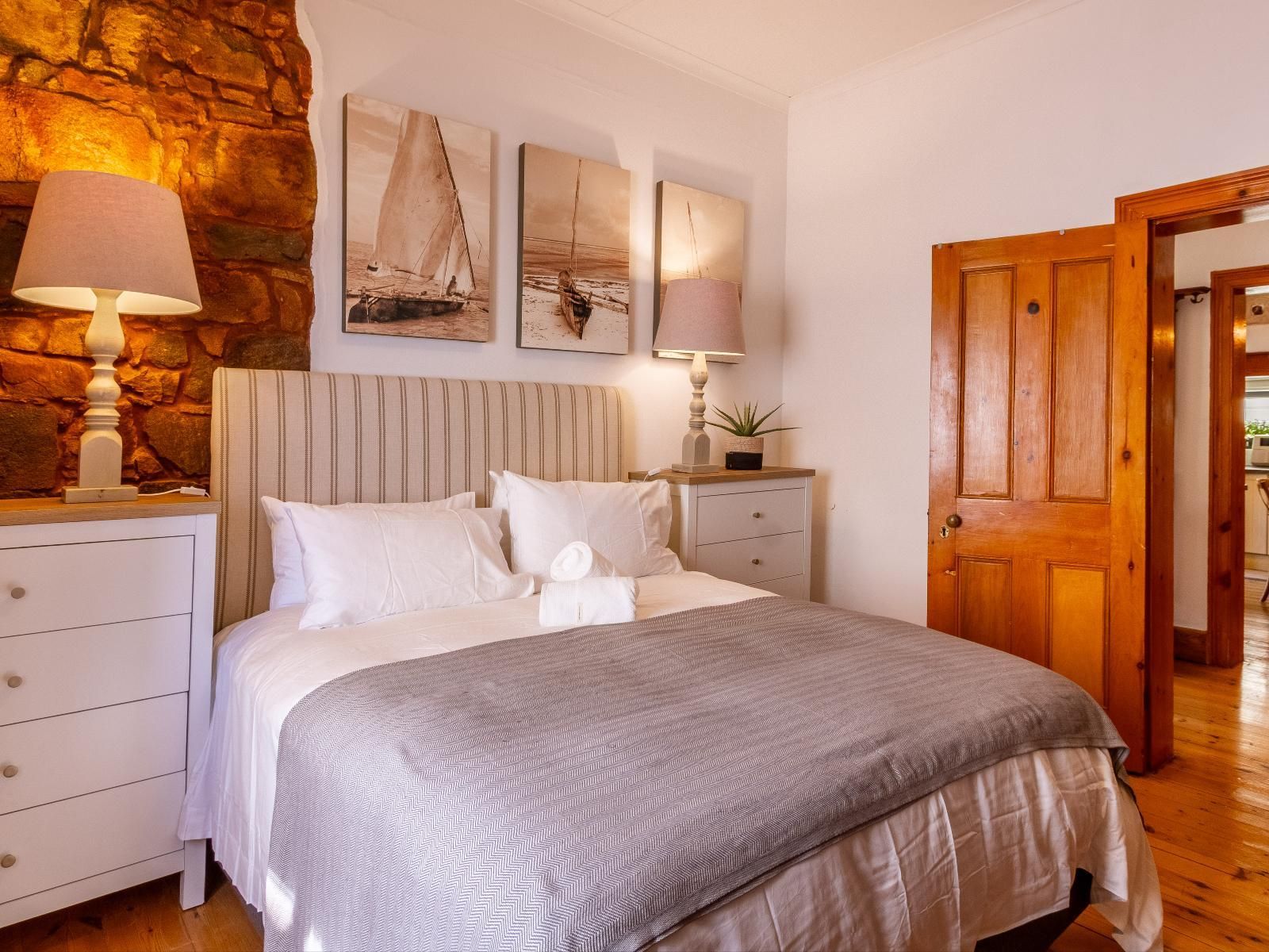 A cozy master bedroom featuring a comfortable bed with white linens and a soft gray throw. The room is warmly lit by bedside lamps on either side of the bed, with a rustic stone wall accent. Nautical-themed artwork hangs above the headboard, adding a coastal touch.