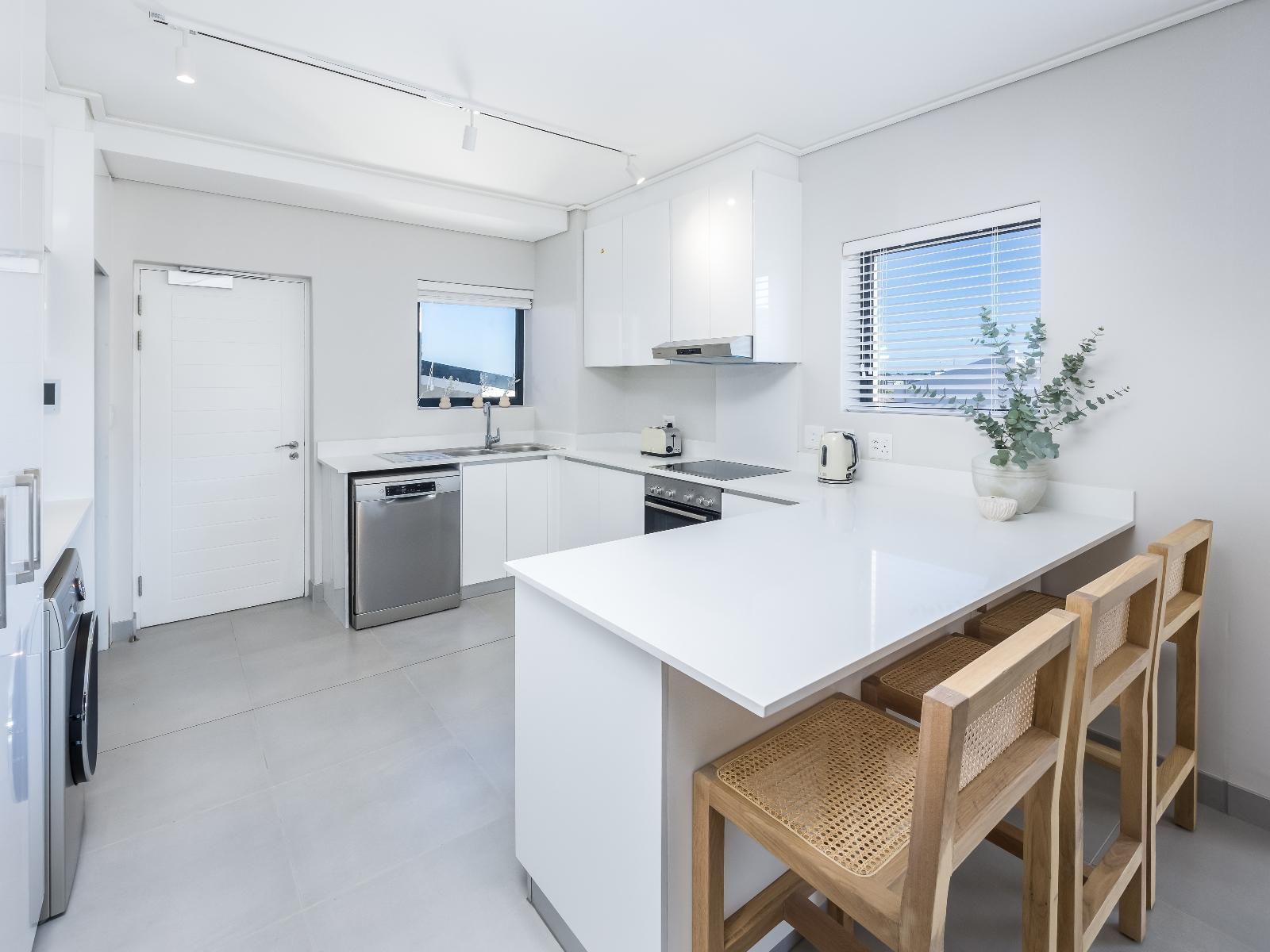 A sleek, minimalist kitchen featuring white cabinetry and countertops. Equipped with a dishwasher, oven, and microwave. Two wooden bar stools sit at the counter. A window provides natural light, complemented by a potted plant for a touch of greenery.