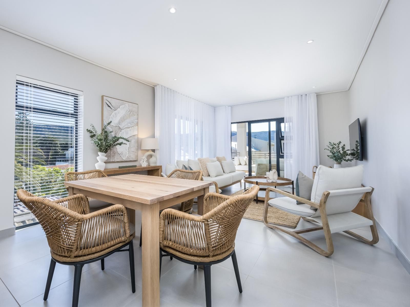 Bright open-plan living room and dining area with wicker chairs and a wooden table. Large windows and a sliding door lead to a private balcony. Neutral tones and minimalist decor create a tranquil atmosphere. The room is equipped with a wall-mounted TV and light furnishings.