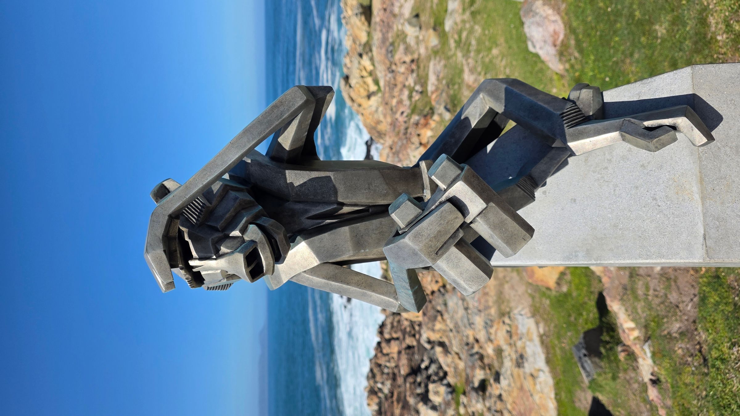 A geometric sculpture of an ape sits on a pedestal against a backdrop of the ocean and rocky shoreline. The metallic figure, crafted with angular shapes, appears to gaze out at the sea, embodying a fusion of modern art and natural beauty in Hermanus.