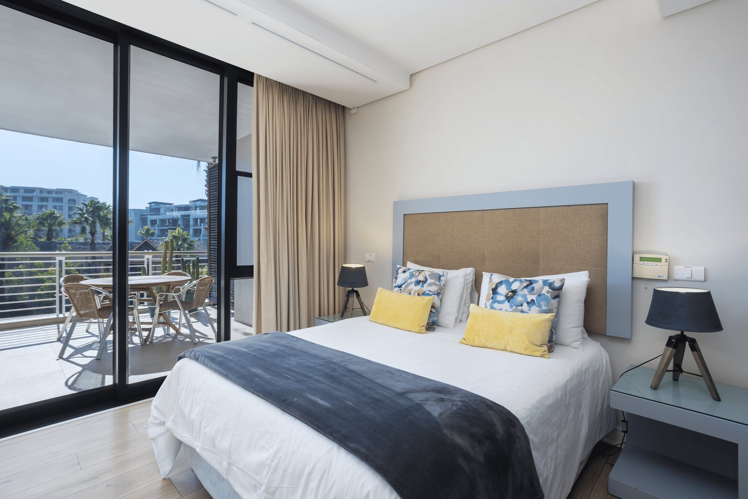 A stylish hotel room featuring a large bed with floral and yellow cushions, flanked by bedside lamps. A sliding glass door reveals a balcony with a table and chairs, overlooking a view of palm trees and modern buildings under a clear blue sky.