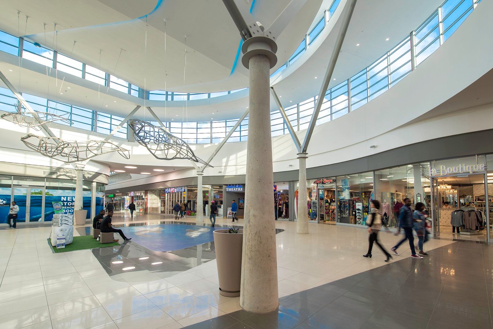 Interior view of Whale Coast Mall featuring modern architecture with high ceilings and large windows. Shoppers walk past various stores, including clothing and specialty shops. The mall is bright and spacious, emphasizing accessibility and a wide range of retail offerings for locals and visitors.
