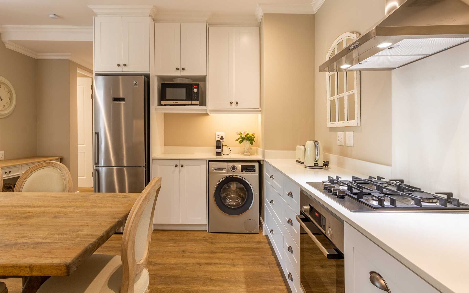 A modern kitchen in Villa 56 featuring white cabinetry, a stainless steel fridge, and a gas stove. It includes a wooden dining table, a microwave, a washing machine, and a toaster, all set against a warm, inviting backdrop. Bright, natural lighting enhances the sophisticated design.
