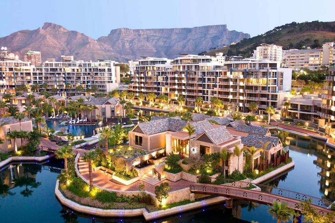 Aerial view of a luxurious resort with modern buildings, lush greenery, and a central waterway. The area is well-lit with pathways and palm trees, set against a backdrop of majestic mountains at dusk.