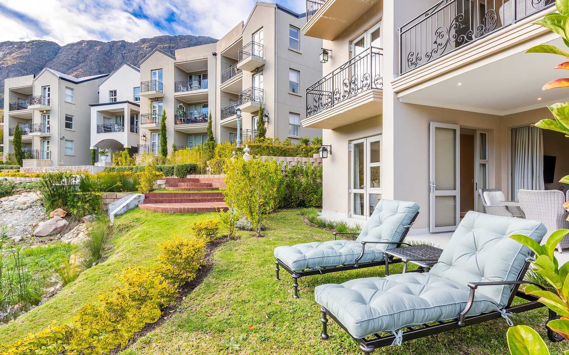 Exterior of a stylish villa with a garden, showcasing two cushioned lounge chairs on a grassy lawn. The villa features balconies with ornate railings and is set against a backdrop of mountains. A small stone path and lush greenery enhance the tranquil atmosphere.