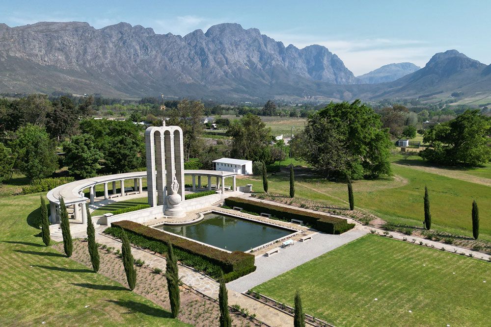 Aerial view of a monument featuring tall columns and a reflecting pool, set in a landscaped garden. The backdrop is a range of rugged mountains under a clear blue sky, surrounded by lush greenery and open fields.